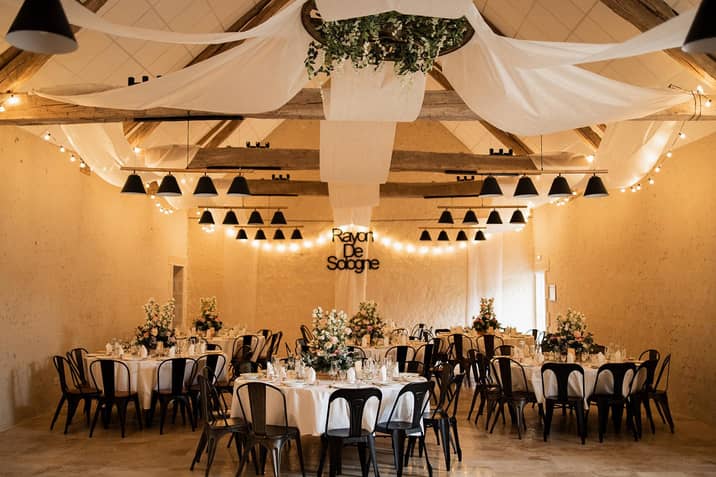 Salle de réception de la grange sous les guirlandes lumineuses, tables rondes dressées